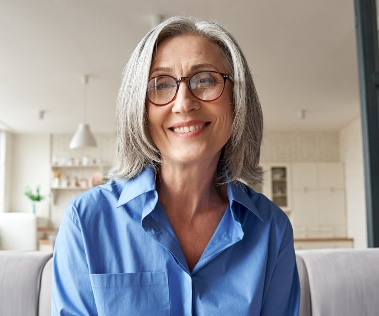 A professional older woman with glasses is dressed in business attire and sitting in her home. She is looking at a laptop camera, smiling.