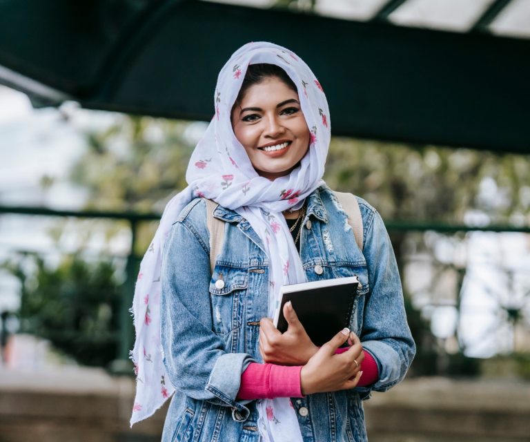 A young woman in a hijab and denim jacket is clasping a notebook to her chest. She is smiling at the camera.