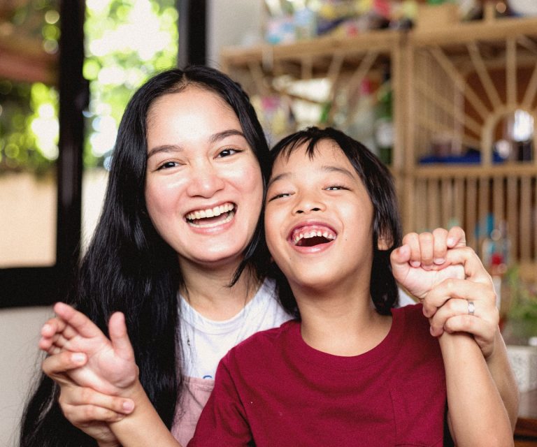 An Asian mother is sitting with her child. She is holding the child's hands. They are both smiling at the camera.